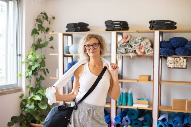 mulher adulta de óculos, na academia, vestindo camiseta regata e calça de moletom, usando mochila na tranversal e segurando uma toalha no pescoço